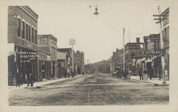 Text on front reads: "Street Scene, Richland Ctr., Wis." Pedestrians are walking on the sidewalks of a main street, with many businesses on each side. A street light is suspended above. Several horse-drawn buggies and wagons are parked on the right. Many businesses have awnings and signs, including, on the left: All America Shoes, Ward Lamberson, and further down the street, Coffland Bros. On the right the business signs are: W. Spickard, Moody & Company Real Estate, Samuel Huffman Flour, Feed & Groceries, and Boston Store. A sign near the roof of a building further down the street reads: "Union Block". Telegraph poles are on the left side of the street. The are hills in the distance.