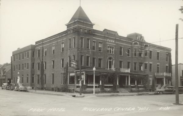 Text on front reads: "Park Hotel, Richland Center, Wis." A large brick hotel is located at the intersection of two city streets. The hotel is three stories tall with porches and balconies. Vines are climbing the columns on the porches. A lamppost is on the corner and automobiles are parked at the curb.