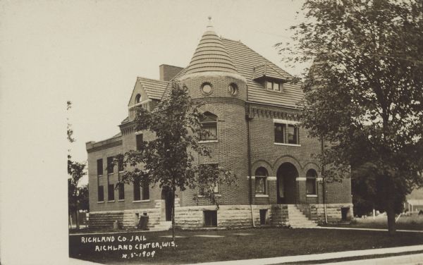 Text on front reads: "Richland Co. Jail, Richland Center, Wis." A large, ornate, brick and stone building surrounded by lawns and sidewalks. There is a tower on the corner of the building and the roof is tiled. A few trees are in the yard and other buildings are in the background.