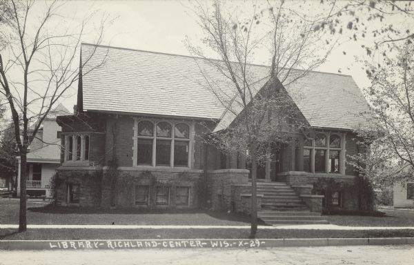 Text on front reads: "Library-Richland Center-Wis." Exterior view of a Carnegie Library built in 1905. The brick and stone building has large windows with arched stained glass panels above. It was located on the southwest corner of East Seminary and Park Streets with sidewalks, lawns and trees. A home is in the background on the left.