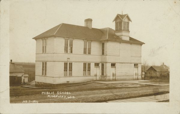Text on front reads: "Public School, Ridgeway, Wis." A two-story clapboard building with a belfry, surrounded by a lawn with sidewalks. Dwellings can be seen on the right and left.