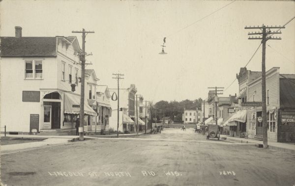 Text on front reads: "Lincoln Street North, Rio, Wis." An unpaved main street with cement sidewalks and crosswalks. There are buildings and storefronts on both sides, many with awnings and signs. Automobiles and horse-drawn vehicles are parked at the curb. Power and telegraph poles line the street and a streetlight is suspended above.