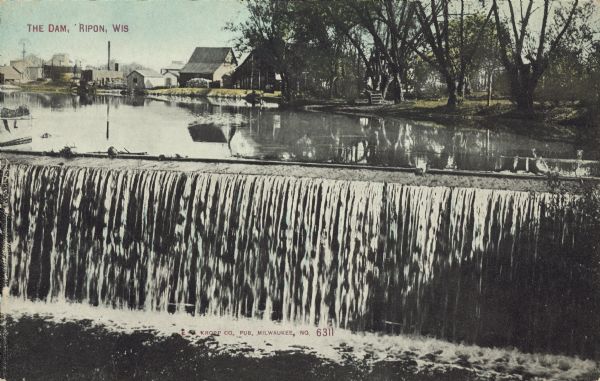 Text on front reads: "The Dam, Ripon, Wis." Water spills over the Ripon Dam, on Silver Creek, with the Ripon Pond in the background. Dwellings and a building can be seen on the shoreline, with trees on the right.