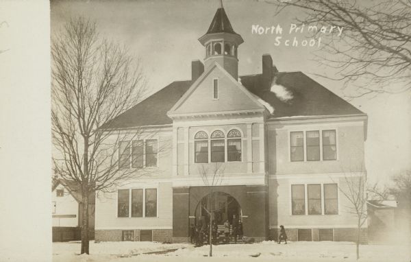 Text on front reads: "North Primary School." A two-story clapboard schoolhouse with a basement has students gathered on the steps to the entrance. The school has a belfry, sidewalks and trees. It is winter and the ground is covered with snow.