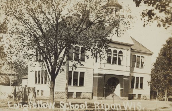 Text on front reads: "Longfellow School, Ripon, Wis." However, this is actually the North Primary School. A two-story clapboard schoolhouse with a basement windows along the foundation, and stairs leading to the entrance. The school has a belfry, sidewalks and trees.