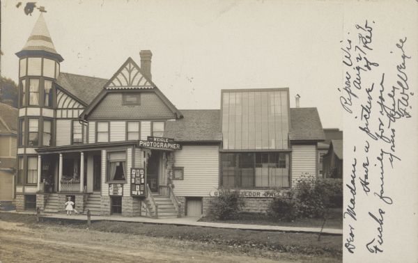 Photographic postcard of a view across a street with a building with signs that read: "Weigle Photographer" and "Ground Floor Gallery". The building is Tudor style with a tower, and the gallery has a two-story window. A little girl is posing on the sidewalk in front of a window. Two boards display photographs from the gallery. A board sidewalk is along the front of the studio and the street is unpaved. There are shrubs in front of the gallery and other buildings are in the background.