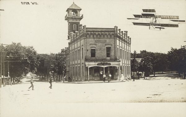 Tall Tale postcard of the City Hall with an airplane added on the right. Text reads: "Ripon, Wis." The City Hall is a brick building with ornate exterior decorations and a bell tower with a balcony, located on the Public Square. There are several pedestrians. Buildings and trees are in the background. The Ripon City Hall building was built in 1900 and removed in 1967. It also housed the post office and fire department.