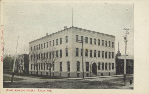 Text on front reads: "Ripon Knitting Works, Ripon, Wis." A large, brick, three-story building with windows to the basement in the foundation. It has an arched entrance, and a sidewalk along the front. The street is unpaved. Other buildings are in the background. 