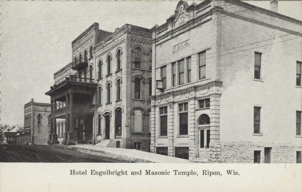 Text on front reads: "Hotel Engelbright and Masonic Temple, Ripon, Wis." The Hotel and Temple stand together on the right side of an unpaved street. There is a sidewalk in front of the buildings. Both buildings have exterior ornamentation and the hotel has a porch and balcony.