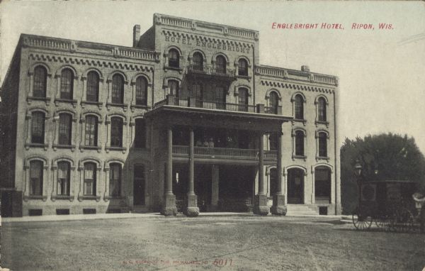 Text on front reads: "Engelbright Hotel, Ripon, Wis." The Hotel stands on an unpaved street with a sidewalk in front. A horse and buggy are on the right. The hotel has exterior ornamentation and several balconies. Several people are standing on the balcony over the entrance.