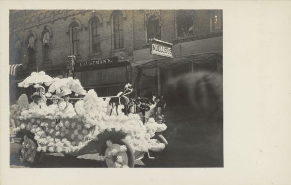 Several people, holding umbrellas, are riding in a car decorated with many tissue paper pom poms for a parade. There are miniature horse models on the hood. The car is driving past brick buildings with patriotic bunting. Two of the businesses are "Faustman's" and "Mueller's".
