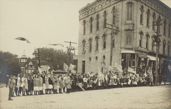 A crowd of adults and children are standing in a line with American Flags on a brick street. There is a large, three-story brick building on the right. In the background on the left is the Weigle Photography Studio.