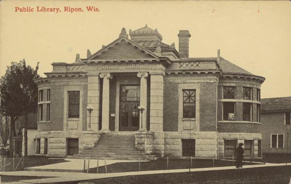 Text on front reads: "Public Library, Ripon, Wis." A one-story brick and stone building with a basement level surrounded by sidewalks. A women is walking along the sidewalk. The Library was opened in 1905 after Andrew Carnegie donated $10,000 to build a new library in 1902. Buildings and trees are in the background.