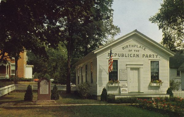 Text on the reverse reads: "The Famous Little White School House, at Ripon, Wisconsin. Birthplace of the Republican Party in 1854." Surrounded by dwellings, trees and a white picket fence, the schoolhouse has signs on it explaining its significance. On the left is a monument. There are flowers along the walk and in boxes at the windows. It has been moved 4 times, the last in 1951. In 1974 it was declared a National Historic Landmark.