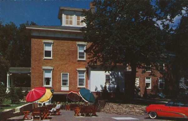 Text on reverse reads: "Stately Republican House Restaurant...at Ripon, Wisconsin, where in 1954, the Republican Party was Born in the 'little White School House,'which is still preserved on the grounds of the Republican House." View of the back of a two-story brick building with a cupola, surrounded by trees. A man is relaxing on a patio with wooden tables, chairs and umbrellas. There is a stone wall behind the patio with an entrance into the basement level. A red automobile is parked on the right.