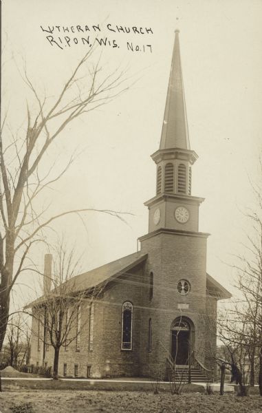 Text on front reads: "Lutheran Church, Ripon, Wis." Front view of a brick church with an arched entrance, steps and a steeple with a clock. A sidewalk runs in front and leafless trees surround the church.