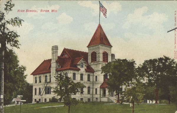 Text on front reads: "High School, Ripon, Wis." A two-story white brick or stone schoolhouse with basement windows along the foundation, and attic space. The school has a belfry with an American flag, chimneys, sidewalks and trees. The roof is tiled in red. 