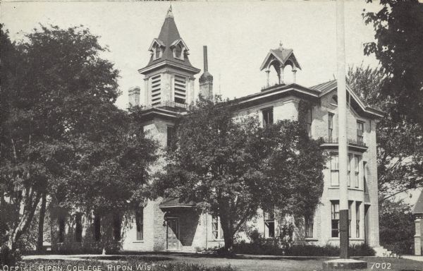 Text on front reads: "Office Ripon College, Ripon Wis." A college building built of stone in 1851 with a steeple and cupola. The building functioned as an office, chapel, music rooms and museum. Mature trees surround the building.