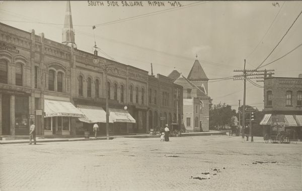 Text on front reads: "South Side Square. Ripon, Wis." View across square towards buildings which line the South Side of the brick paved Public Square. Several building have awnings with store names on them. In the center, a woman and a child are walking on the square, on the left is a man. Another man is on the sidewalk. Several horse-drawn vehicles are on the right.