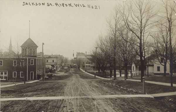 Text on front reads: "Jackson St., Ripon, Wis." A view down an unpaved street, sidewalks, buildings and dwellings on both sides. Many trees can be seen, mostly on the right.