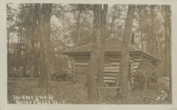 Text on front reads: "In the Park, River Falls, Wis." A log cabin or shelter surrounded by picnic tables and many mature trees.
