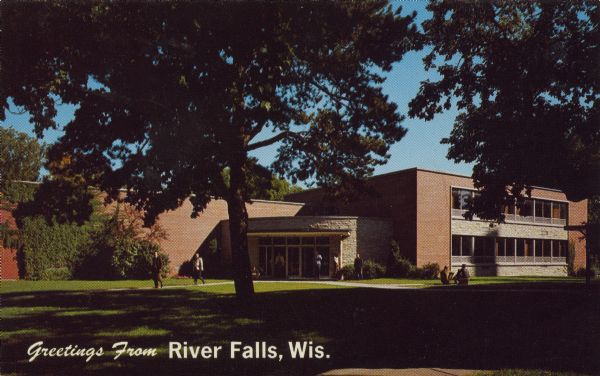 Text on front reads: "Greetings from River Falls, Wis." Text on the reverse reads: "The Chalmer Davee Library was built in 1954. In addition to the usual facilities it houses the Little Theatre of the Wisconsin State University at River Falls." A red brick building with stone accents seen through mature trees. Several people can be seen leaving the building and at a patio table. The university is now called UW-River Falls.
