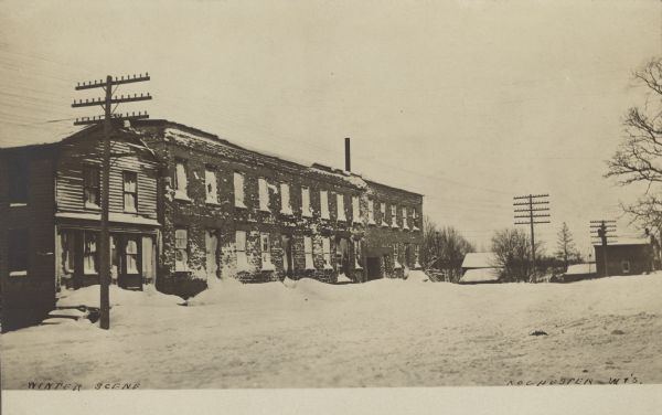 Text on front reads: "Winter Scene, Rochester, Wis." Snow covers a long, two-story brick building and dwellings in a small town. Snow is mounded up against the foundations.