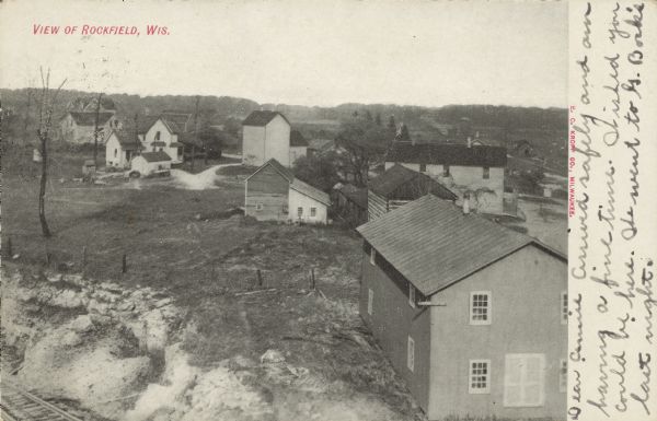 Text on front reads: "View of Rockfield, Wis." Elevated view of a small town, with buildings, dwellings and a log cabin . Railroad tracks are in the lower left corner.