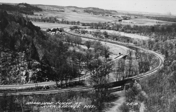 Text on front reads: "Horseshoe Curve At Rock Springs, Wis." A road and a railroad track, with bridges over the Baraboo River, make a double curve in an elevated view of the Upper Narrows. Text on reverse reads: "Rock Springs, Wis., Noted for Artesian Springs Amid Scenic Rock Gorges. A scene in the geologically famous UPPER NARROWS of the Baraboo Quartzite Range, located at Rock Springs, on scenic state highway 136, attracting visitors from far and wide. Only 30 minutes drive from Wisconsin Dells or Devil's Lake, Wis."