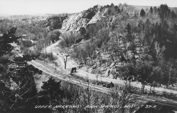 Text on front reads: "Upper Narrows, Rock Springs, Wis." A road and a railroad tracks run next to each other in an elevated view of the Upper Narrows. Text on reverse reads: "Rock Springs, Wis., Noted for Artesian Springs Amid Scenic Rock Gorges. A scene in the geologically famous UPPER NARROWS of the Baraboo Quartzite Range, located at Rock Springs, on scenic state highway 136, attracting visitors from far and wide. Only 30 minutes drive from Wisconsin Dells or Devil's Lake, Wis."