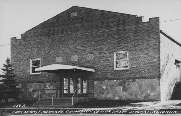 Text on front reads: "Rock Springs Memorial Community Center." The front view of the Community Center built of brick and stone. Text on reverse reads: "Rock Springs Memorial Center. Built by community effort and a spirit of Co-operation by the people of Rock Springs, Wis. and surrounding area." Built in 1950, the Community Center was severely damaged in a flood in 2018 and plans are underway to rebuild in a new location.