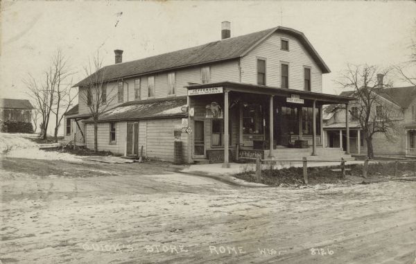 Text on front reads: "Quick's Store, Rome, Wis." A general store in a small town with a porch along the front. the signs read: "Jefferson, Wagons and Implements", "Rome, Post Office", "Polarine," "Red Crown Gasoline" and "Telephone". The street is unpaved with concrete sidewalks.