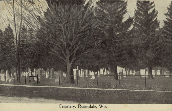Text on front reads: "Cemetery, Rosendale, Wis." A cemetery inside a chain link fence with mature trees. A sidewalk runs along the unpaved road in the foreground.