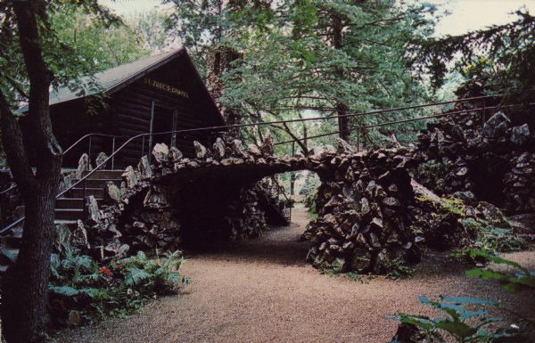 Text on back reads: "Log Chapel dedicated to St. Jude, Apostle, at the terminus of a bridge from which a wayside shrine and World War-I Memorial is viewed. Grotto Shrine & Wonder Cave. St. Philip the Apostle Church. Rudolph, WI. 54475. Shrine dedicated to 'Peace' by its founder Father Philip J. Wagner (1918-1959)." A Chapel made of logs with a bridge and walls built of stones. Trees surround the Chapel.