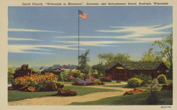Text on front reads: "Small Church, 'Wisconsin in Miniature,' Souvenir and Refreshment Stand, Rudolph, Wisconsin." A garden with flowers, rocks, shrubs, paths, a lawn and an American flag. In the background are log buildings and trees.