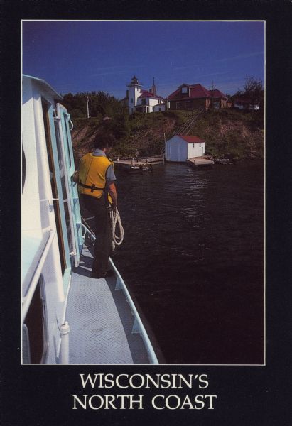 Text on front reads: "Wisconsin's North Coast." A man is standing on the side deck of a boat, and in the distance is the Raspberry Island Lighthouse with a dock below. Text on the reverse reads: "Explore Wisconsin. Raspberry Island, Lake Superior - Climb the historic Raspberry Island Lighthouse on an Apostle Islands Cruise! Or explore Wisconsin's other national treasures - the Nicolet and Chequamegon National Forests, and the St. Croix National Scenic Riverway. For a free Wisconsin Vacation Kit, write: Wisconsin Dept. of Development - Tourism, Box 7606, Madison, WI 53707, or call toll-free from Wisconsin or adjoining states 1-800-ESCAPES."