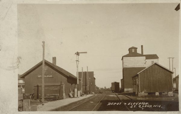 Text on front reads: "Depot and Elevator, St. Cloud, Wis." A view up the railroad tracks with the station on the left and grain elevator on the right, with other industrial buildings. A grain car is on the tracks.
