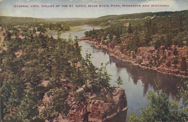 Text on front reads: "Dalles of the St. Croix, Inter State Park, Minnesota and Wisconsin." Elevated view of the St. Croix River showing the rock formations on the shorelines, an automobile bridge and a footbridge on the lower left. The buildings of a town can be seen on the upper left and trees cover the land. 