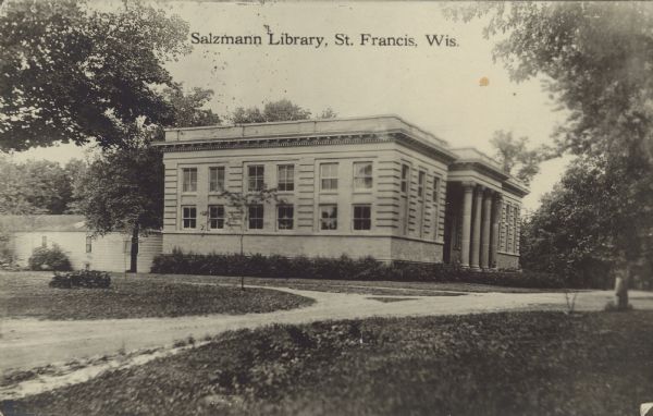 Text on front reads: "Salzmann Library, St. Francis, Wis." A stone and brick building with columns at the entrance. An unpaved street runs past it and trees are in the background. It is one of the oldest seminary libraries in the United States.
