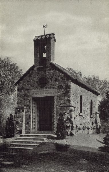 A stone Chapel, St. Mary of the Angels has an ornate entrance and a belfry. Decorated steps, a lawn and trees can be seen.