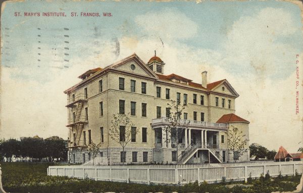 Text on front reads: "St. Mary's Institute, St. Francis, Wis." A Catholic School for Girls opened in 1904. It is a three-story building on an elevated foundation built of Milwaukee Cream City bricks. The entrance has a porch with a double staircase. The yard is enclosed with a white picket fence and trees are on the horizon.