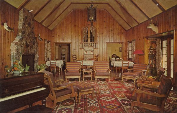 Text on reverse reads: "Lofty Pines Lodge. Big St. Germain Lake. St. Germain, Wisconsin. American and European Plan." Interior view of a lodge at a Northern Wisconsin resort, with knotty pine paneling and a stone fireplace. It is furnished with chairs, tables, a piano, and a patterned rug. There is an elaborate chandelier and taxidermy on the walls. 