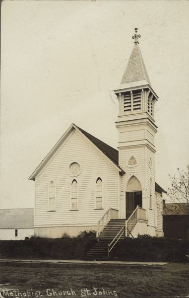 Caption reads: "Methodist Church, St. Johns." A clapboard church with a steeple. There are steps from the sidewalk to the entrance and the street is unpaved.