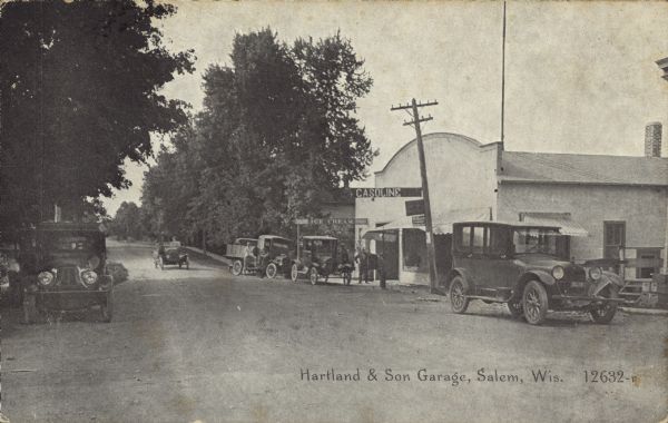 Text on front reads: "Hartland & Son Garage, Salem, Wis." A building with an arched roof and square false corners on the front. Many automobiles and trucks are parked at the curb, and one auto is in the street. Several people are standing in front, under sign that reads: "Gasoline." Many trees line the street.