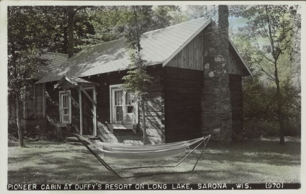 Text on front reads: "Pioneer Cabin at Duffy's Resort on Long Lake, Sarona, Wis." A square hewn log cabin at a resort has a stone chimney and is surrounded with trees. A hammock is on the front lawn. Colorized photograph.
