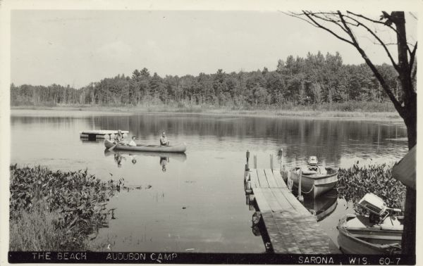 Text on front reads: "The Beach, Audubon Camp. Sarona, Wis." A view of a wooden dock and lake from the shore. Three people are in a canoe, with a floating raft behind them. Two power boats are tied up at the dock. Trees are along the far shoreline.