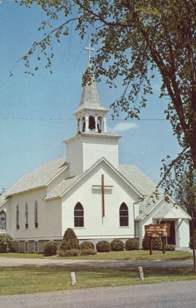 Text on reverse reads: "Long Lake Lutheran Church (ALC). Route 1, Sarona Wis. 54870. In Washburn County, on county Trunk D, East of Highway M, Sound end of Long Lake. Services year 'round 11:00 A.M. Summer Schedule: 8:00 A.M. Outdoor Service, Weather Permitting, 11:00 A.M. at Church." A white clapboard church with a steeple and bell. A street and driveway are in the foreground, with a tall tree on the right and shrubs around the foundation.