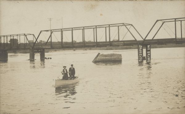 Text handwritten on bridge: "Toll Bridge, Sauk City." A couple are standing in a canoe on the Wisconsin River, in front of the Toll Bridge. Old supports can be seen on the other side of the bridge and the shoreline is lined with trees.