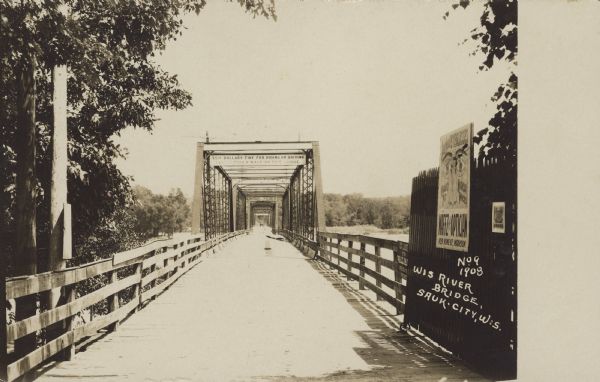 Text handwritten on right: "Wis. River Bridge, Sauk City, Wis." A view straight through the bridge girders to the opposite side. Signs can be seen on the open gate. There are trees on the near side and covering the far shoreline.