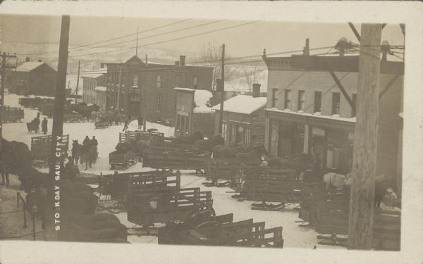Text at left reads: "Stock Day, Sauk City." Horse-drawn sleds are hitched on both sides of a snow covered street lined with businesses. Townspeople are on foot or driving their sleds. Some of the storefront signs read: "Millinery," "Boots and Shoes" and "Eagle House, Math. Huerth."